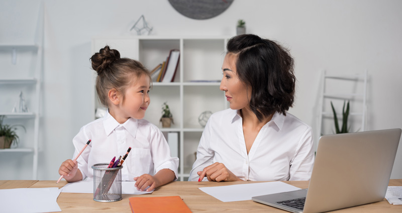 Asian Mother And Daughter Sitting And Looking At Each Other At Desk With Pens Paper And Laptop