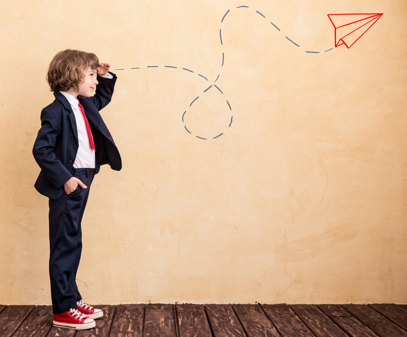 Young Boy In Business Suit With Hand Over Eyes Looking Into Distance With Drawing of Paper Plane On Wall