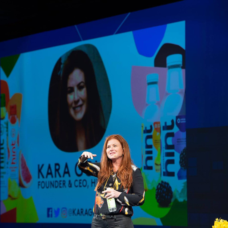 Kara Goldin On Stage Delivering Speech With Hint Water Bottle In Her Hand And Large Screen In Background