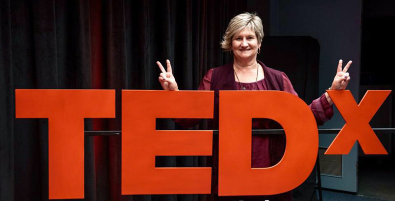 Kelly Garrett Posing In Front Of TEDx Sign Giving Peace Sign With Fingers