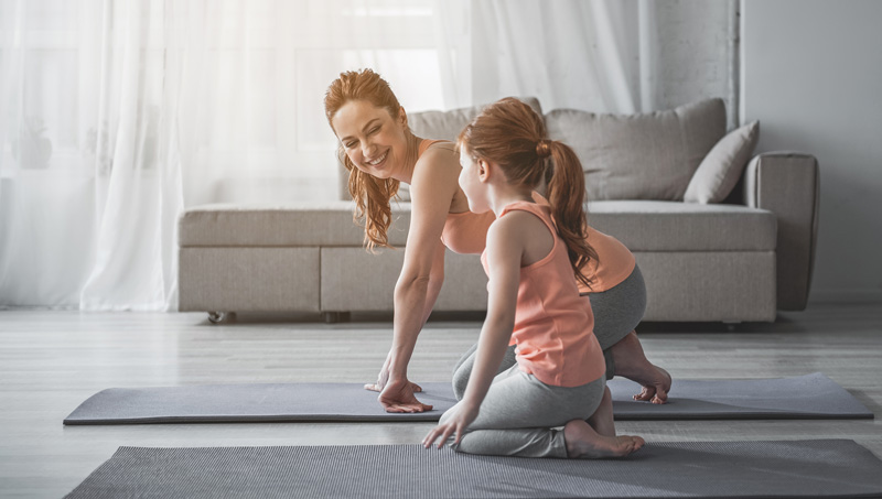 Mother Smiling To Daughter While Doing Yoga Together In Living Room